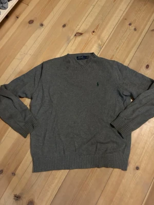 Grå tröja från Polo Ralph Lauren - Klassisk grå tröja från Polo Ralph Lauren med rund halsringning och diskret broderad logga på bröstet. Tröjan har ribbade muddar vid ärmslut och nederkant, perfekt för en stilren och enkel look.