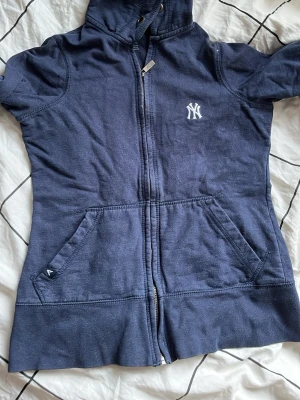 Mörkblå New York Yankees hoodie - Mörkblå hoodie med dragkedja från New York Yankees. Klassisk logga på bröstet, två stora fickor framtill och justerbar huva med snören. Mjuk bomullskänsla och sportig look, perfekt för en avslappnad stil.