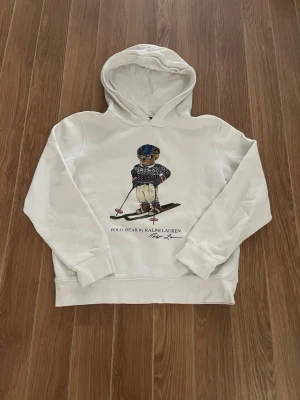 Vit hoodie Polo Bear Ralph Lauren - Vit hoodie från Ralph Lauren med Polo Bear-tryck där björnen åker skidor. Klassisk huva och ribbade muddar. Snygg och trendig design med broderad detalj framtill. Perfekt för dig som gillar streetwear och ikoniska motiv. Storlek XS L (14-16)