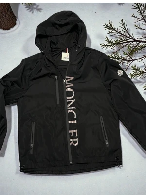 Svart vindjacka från Moncler - Oanvänd  svart vindjacka från Moncler med stor logga framtill. Jackan har huva, tre dragkedjeförsedda fickor och Moncler-märke på ärmen. Tillverkad i lätt, vattenavvisande material som passar perfekt för blåsiga dagar.