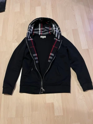 Svart hoodie från Burberry med dragkedja - Säljer en svart hoodie från Burberry med klassiskt rutigt foder i huvan. Hoodien har dragkedja framtill, två fickor och justerbar dragsko i huvan. Tillverkad i mjukt bomullsmaterial och har diskret Burberry-logga på bröstet. Perfekt för en stilren och avslappnad look. Kedjan knasar ibland men annars funkar bra när man haft på sig den 