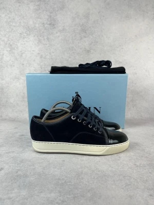 Marinblå sneakers från Lanvin - Snygga marinblå sneakers från Lanvin med ovandel i mocka och lackad tå i svart. Skorna har vita sulor, mörkblå skosnören och silverfärgade öljetter. Perfekta för dig som vill ha en clean och stilren look.