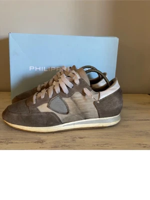 Philippe Model beige/bruna sneakers - Snygga sneakers från Philippe Model i beige och brunt med detaljer i mocka och textil. Skorna har vita skosnören, låg profil och klassisk design med märkets logga på sidan. Perfekta för dig som gillar stilrena och trendiga sneakers.