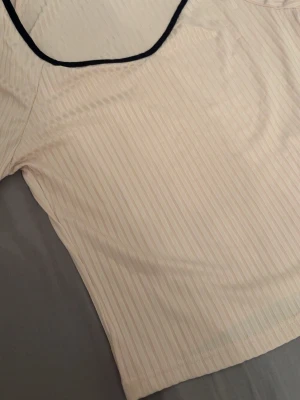 Beige ribbad topp från SHEIN - Säljer en beige ribbad topp från SHEIN i storlek M. Toppen har svarta kanter vid halsringningen och ett diskret randigt mönster i materialet. Perfekt för dig som gillar enkel men snygg stil.