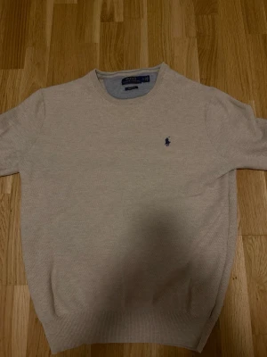 Beige stickad tröja från Polo Ralph Lauren - Snygg beige stickad tröja från Polo Ralph Lauren med klassisk rund halsringning och den ikoniska mörkblå broderade loggan på bröstet. Tröjan har ribbade muddar vid ärmslut och nederkant. Perfekt för en stilren och avslappnad look.