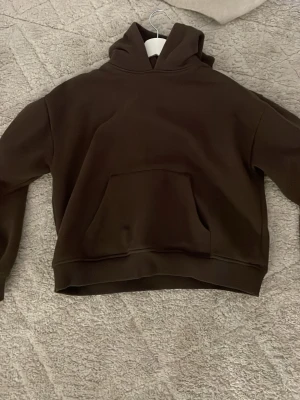 Mörkbrun hoodie med magficka - Snygg mörkbrun hoodie med klassisk huva och stor magficka framtill. Hoodien har en avslappnad passform och ribbade muddar vid ärmar och nederkant. Perfekt för en chill och casual look. Materialet känns mjukt och bekvämt. Jätte fint skick