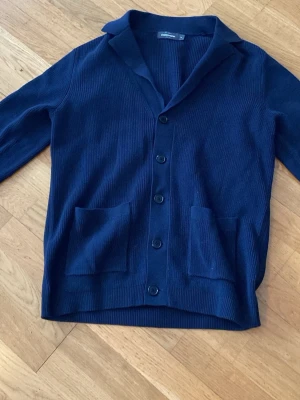 Mörkblå kofta från Dressmann - Stilren mörkblå kofta från Dressmann med ribbad struktur, knäppning framtill och två stora fickor. Klassisk krage och lång ärm. Perfekt för lager på lager och enkel att matcha med olika outfits. Skriv för mått 