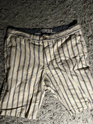 Randiga linne shorts från Dressmann - Säljer ett par beige linne shorts från Dressmann med blå och vita vertikala linjer. Bra skick och har använts fåtal gånger. Säljes för jag inte använder de längre. Köptes för 399kr och säljes nu för 150kr. Kontakta mig om du har några frågor.