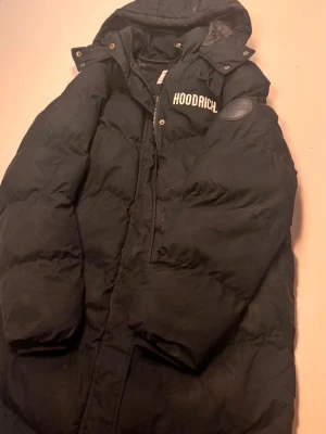 Svart dunjacka från Hoodrich - Snygg svart dunjacka från Hoodrich med logga på bröstet och ett runt emblem på ärmen. Jackan har huva med tryckknappar och dragkedja framtill. Perfekt för kalla dagar och ger en streetwear-vibe. Vill sälja den eftersom den är inte min stil nu. Jag har använt den bara den hära vinten. Alltså nästan i 2 månader. Man blir inte kall upp till -35 grader kallt jag har provat i Kiruna och frös inte.