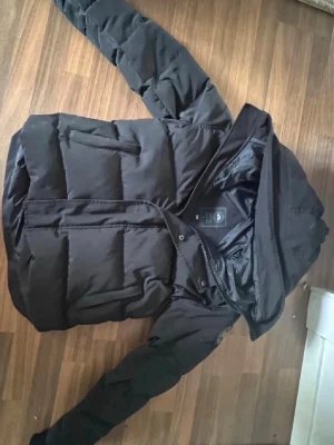 Canada goose - Snygg svart pufferjacka med huva och dragkedja framtill. Jackan har en quiltad design med breda paneler och är tillverkad i ett glansigt material som ger en modern look. Perfekt för kalla dagar och enkel att matcha med olika outfits.
