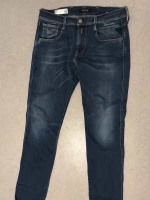 Mörkblå slim fit jeans från Replay - Säljer ett par mörkblå slim fit jeans från Replay med snygga slitningar och kontrastsömmar. Jeansen har klassiska fem fickor och en något tvättad look framtill och baktill. Perfekta för dig som gillar en modern och avslappnad stil.
