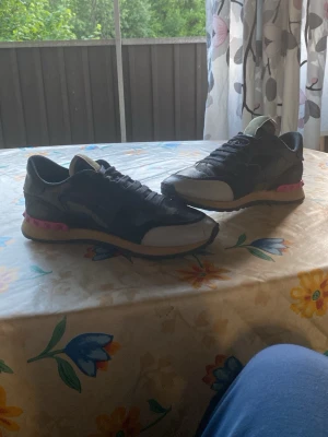 Svarta sneakers med camomönster - Säljer ett par svarta sneakers med camomönster och vita detaljer. Skorna har svarta skosnören, beige sula med rosa inslag och en klassisk låg siluett. Materialet verkar vara en mix av mocka och textil. Perfekta för dig som gillar streetwear och vill sticka ut lite extra.