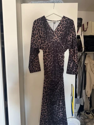 Lång leopardmönstrad klänning - Snygg långklänning med leopardmönster och v-ringning. Klänningen har långa ärmar och markerad midja som ger en smickrande siluett. Perfekt för dig som vill sticka ut med ett djurmönstrat plagg.