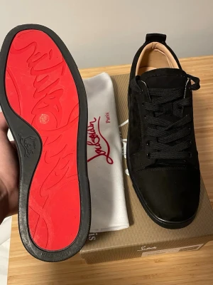 Svarta sneakers från Christian Louboutin - Snygga svarta sneakers från Christian Louboutin med klassisk röd sula och diskret logga. Skorna har ovandel i mocka, svarta skosnören och en låg profil. Perfekta för dig som vill ha en stilren och exklusiv look.