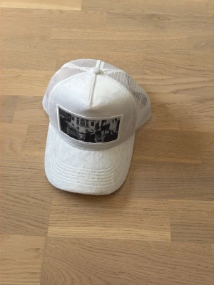 Vit meshkeps med svartvit patch - Säljer en vit keps med mesh på sidorna och bak, samt en svartvit patch med motiv framtill. Kepsen har böjd skärm och justerbar baksida. Från Emilio Pendenza