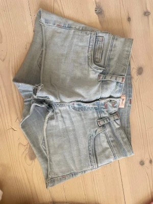 Ljusblå jeansshorts med broderi - Säljer ett par ljusblå jeansshorts från Replay med råa kanter och röda broderade detaljer på bakfickorna. Klassisk femficksmodell med bälteshällor och knappgylf. Perfekta för varma dagar och enkel att matcha med olika toppar.