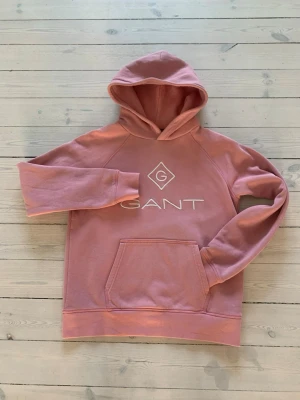 Rosa hoodie från GANT med logga - Säljer en rosa hoodie från GANT med stor vit logga och diamantmönster på bröstet. Hoodien har en klassisk känguruficka framtill, huva och ribbade muddar. Perfekt för dig som gillar en avslappnad och snygg stil. Materialet känns mjukt och skönt.