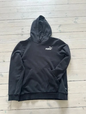 Svart hoodie från Puma med logga - Svart hoodie från Puma med vit logga på bröstet. Klassisk modell med huva och känguruficka framtill. Tillverkad i mjukt material som känns skönt mot huden. Perfekt för en avslappnad och sportig stil.
