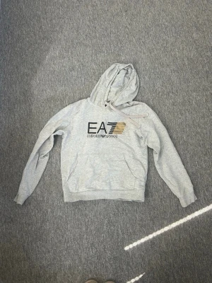 Grå hoodie från EA7 Emporio Armani - Snygg ljusgrå hoodie från EA7 Emporio Armani med stor logga i svart och guld på bröstet. Använd o fått lite missfärgning i tvätten.