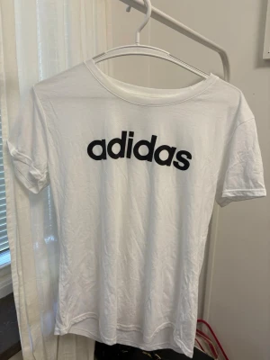 Vit Adidas t-shirt med logga - Vit t-shirt från Adidas med svart logga tryckt framtill. Klassisk rund halsringning och korta ärmar. Tillverkad i en mjuk mix av polyester, bomull och viskos som ger en skön känsla och bra passform.