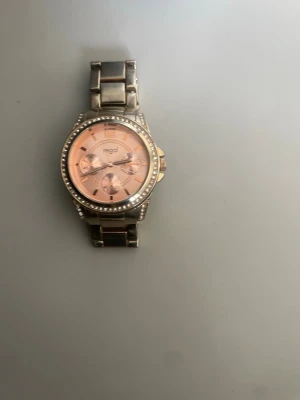 Regal klocka med rosa urtavla - Snygg klocka från Regal med metallarmband och rosa urtavla. Runt boetten sitter små glittrande stenar som ger extra bling. Klockan har flera dekorativa urtavlor och en klassisk, rund form. Perfekt accessoar för att lyfta din stil.