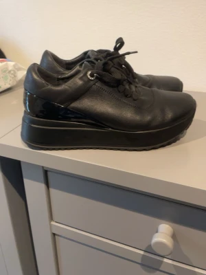 Svarta platåsneakers i skinn - Snygga svarta sneakers med platåsula och snörning. Tillverkade i slätt skinn med detaljer i blankt material vid hälen. Tjock, räfflad gummisula för extra höjd och cool look. Perfekta för dig som vill ha lite extra edge i vardagen.