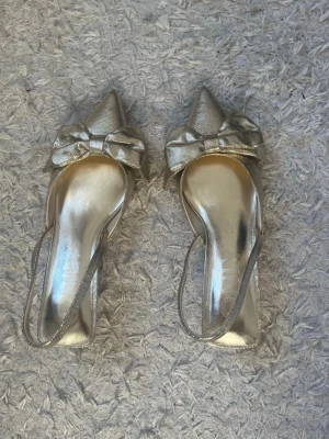 Guldiga pumps med rosett från ONLY - Snygga guldiga pumps från ONLY med spetsig tå, låg klack och öppen häl med rem. Dekorativa stora rosetter framtill ger en extra glamorös touch. Perfekta för dig som vill sticka ut och addera lite bling till din outfit. 4,5 cm klack. Använda en gång så lite smutsiga i ytter sulan