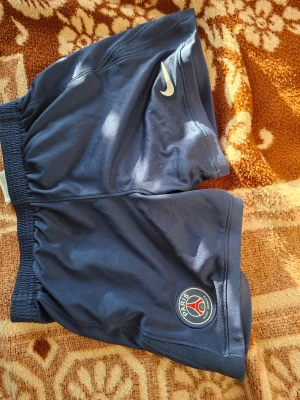 PSG matchställ Nike blå/röd - Säljer ett blått PSG matchställ från Nike med tröja och shorts. Tröjan har röd och vit rand i mitten, PSG-logga och Qatar Airways-tryck. Shortsen har PSG-logga och Nike Swoosh. Tillverkat i lätt och ventilerande material, perfekt för fotbollsträning.