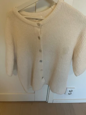 Vit stickad kofta med knappar - Mysig vit stickad kofta med rund hals och knäppning framtill. Koftan har en mjuk, fluffig känsla och ribbad struktur. Perfekt att slänga över en t-shirt eller linne för en avslappnad look.
