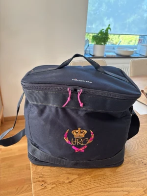 Svart ryggsäck från Fouganza - Rymlig svart ryggsäck från Fouganza med broderad krona och texten HRC på framsidan. Väskan har rosa dragkedjor, handtag upptill och justerbar axelrem. Insidan är fodrad i stark rosa färg och har nätficka med dragkedja. Väskan kan säljas enskild men helst med all häst redskap!