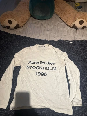 Vit långärmad topp Acne Studios - Vit långärmad topp från Acne Studios med tryck i svart text på bröstet där det står 'Acne Studios STOCKHOLM 1996'. Klassisk rund halsringning och rak passform. Perfekt för dig som gillar minimalistisk och stilren design.