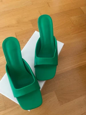 Gröna slip-in pumps med fyrkantig tå - Snygga gröna slip-in pumps med öppen häl och fyrkantig tå. Skorna har en bred rem över foten och smal klack. Tillverkade i ett slätt material som ger en modern look. Perfekta för dig som vill sticka ut med färg och stil.