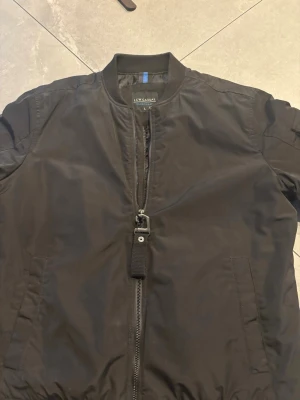 Svart bomberjacka från LCW Casual - Snygg svart bomberjacka från LCW Casual med klassisk ribbad krage och muddar. Jackan har dragkedja framtill, två sidofickor och en detalj med band vid dragkedjan. Tillverkad i ett glansigt syntetmaterial som ger en modern look.