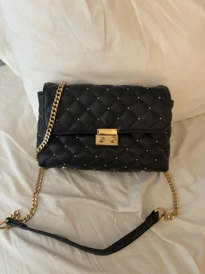 Svart quiltad handväska från Cortefiel - Snygg svart quiltad handväska från Cortefiel med guldfärgade nitar och kedjerem. Väskan har ett elegant lås framtill och innerfack med dragkedja. Perfekt accessoar för dig som gillar en klassisk men trendig look.