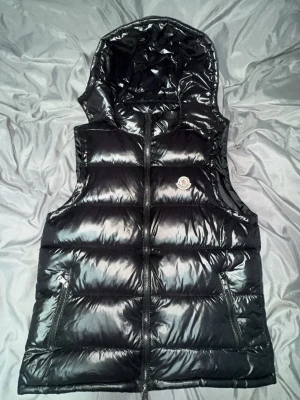 Svart dunväst från Moncler med huva - Snygg svart dunväst från Moncler med glansig finish och avtagbar huva. Västen har två dragkedjefickor framtill, quiltad design och klassisk Moncler-logga på bröstet. Perfekt för lager-på-lager och streetstyle. Ser mycket bättre ut i verkligheten! 