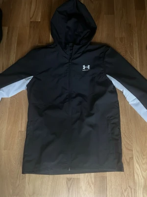 Svart vindjacka från Under Armour - Sprillans ny snygg svart vindjacka från Under Armour med vit logga på bröstet och vita detaljer längs ärmarna. Jackan har huva, dragkedja framtill och meshfoder på insidan för extra komfort. Perfekt för blåsiga dagar och sportiga outfits.