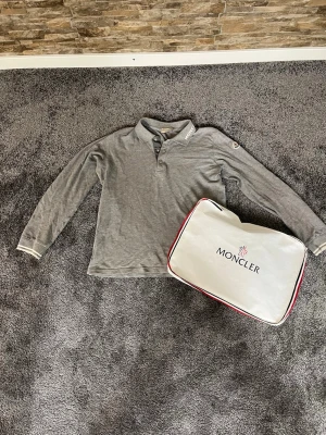 Grå Moncler pikétröja långärmad - Säljer en stilren grå långärmad pikétröja från Moncler med klassisk krage och tre knappar framtill. Moncler-logga syns på kragen och ärmen. Perfekt för dig som gillar exklusiva märken och enkel design.