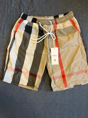 Randiga shorts med snörning - Snygga shorts med breda ränder i beige, svart, rött och vitt. De har elastisk midja med vit snörning och är tillverkade i ett lätt syntetmaterial. Perfekta för sommaren och har en modern, avslappnad look.