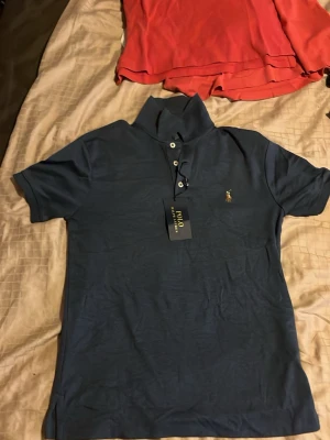 Blå pikétröja från Polo Ralph Lauren - Snygg mörkblå pikétröja från Polo Ralph Lauren med klassisk krage, korta ärmar och knappar framtill. Tröjan har den ikoniska broderade loggan på bröstet och är tillverkad i mjuk bomull som känns skön mot huden. Den är 100% äkta och har inte använts nån gång, sjukt bra kvalité. Inget annat än ett klipp inför sommaren när den säljes för 349kr och kostar 995kr. Säljer för endast 349kr eftersom jag vill få något för den eftersom den inte kommer till användning och vill få någonting för den.