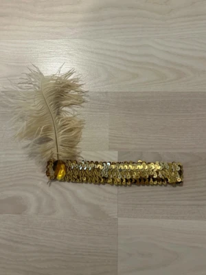 Guldigt diadem med paljetter och fjäder - Säljer ett glittrigt diadem täckt av guldiga paljetter och dekorerat med en stor beige fjäder på sidan. Perfekt för dig som vill sticka ut med en vintageinspirerad accessoar. Diademet har elastiskt band och passar de flesta huvudstorlekar.