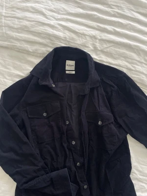 Svart overshirt från Dobber - Stilren svart overshirt från Dobber i manchestertyg. Skjortan har två bröstfickor med lock och knappar, klassisk krage och knäppning framtill. Perfekt att bära öppen över en t-shirt för en avslappnad look.