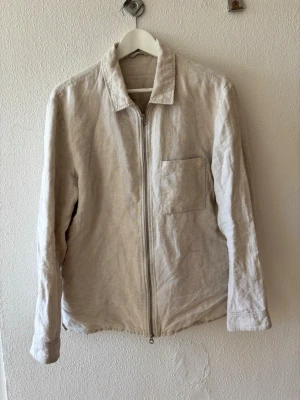 Beige linneskjorta overshirt med dragkedja - Stilren overshirt i beige linne från J.Lindeberg  med dragkedja framtill och en bröstficka. Klassisk krage och avslappnad passform, perfekt för lager på lager. Materialet är luftigt och ger en naturlig look.