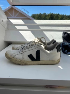 Vita Veja sneakers med svart detalj - Snygga vita Veja sneakers med svart V-logga på sidan och svart hälflik med VEJA-tryck. Klassisk låg modell med vita snören och gummisula. Tillverkade i läder och passar perfekt till jeans eller kjol.