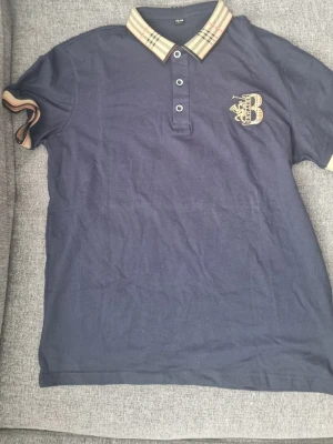 Marinblå piké från Best Men - Snygg marinblå pikétröja från Best Men med klassisk krage i beige och rutigt mönster. Broderad logga med bokstaven B och texten Best Men på bröstet. Kortärmad modell i mjuk bomull, perfekt till en avslappnad stil. Det står storlek L men känns mer som S till XS