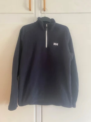 Svart fleece från Helly Hansen - Mysig svart fleece från Helly Hansen med halv dragkedja och hög krage. Tröjan har diskret HH-logga på bröstet och är perfekt för lager-på-lager. Långärmad och i mjukt material som håller dig varm under kyliga dagar.