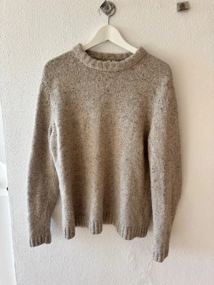 Beige stickad tröja med rund hals från arket  - Mysig beige stickad tröja från Arket med rund hals och ribbade muddar vid ärmslut och nederkant. Tröjan har en enkel, rak passform och är tillverkad i ett mjukt, melerat garn som ger en avslappnad look. Perfekt för lager på lager under kyliga dagar.