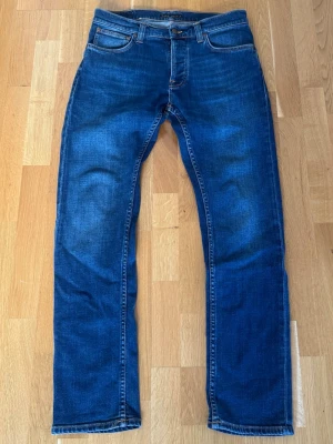 Nudie Jeans Grim Tim  - Säljer ett par blåa jeans från Nudie Jeans med klassisk slim passform. Jeansen har orangea sömmar och Nudie-logga på bakfickan. Tillverkade i slitstark denim med snygg tvätt och subtila slitningar. Dessa är i toppppp skick knappast använda, modell Grim Tim, sitter som W31