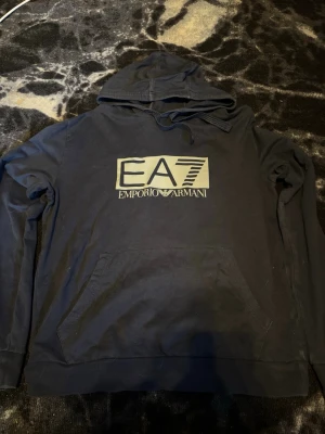 Svart hoodie från Emporio Armani EA7 - Säljer en svart hoodie från Emporio Armani EA7 med stor logga framtill. Hoodien har huva med dragsko och en klassisk känguruficka. Tillverkad i mjukt bomullsmaterial och har långa ärmar. Perfekt för dig som gillar stilrena och trendiga plagg. Tvättar innan jag skickar iväg ☺️
