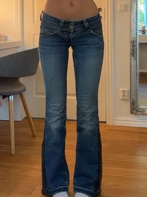 Blå bootcut jeans med låg midja - Snygga blå bootcut jeans med låg midja och dubbla knappar framtill. Jeansen har klassiska fickor. Perfekta för dig som gillar ett par jeans som sticker ut.