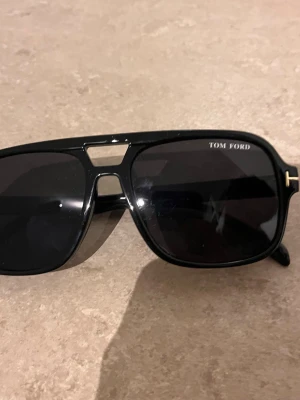 Svarta solglasögon från Tom Ford - Stilrena svarta solglasögon från Tom Ford med markerad båge och mörka glas. Klassisk pilotform med snygga gulddetaljer vid skalmarna. Perfekt accessoar för att lyfta din look.